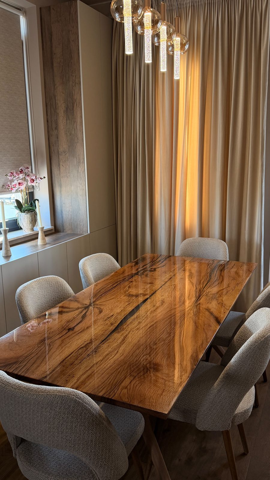 Solid oak dining table with a resin-filled natural crack and high-gloss epoxy finish under glass pendant lights, surrounded by six beige bouclé chairs